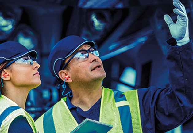 Two people in bump caps, safety glasses and ear protection look up at an engine